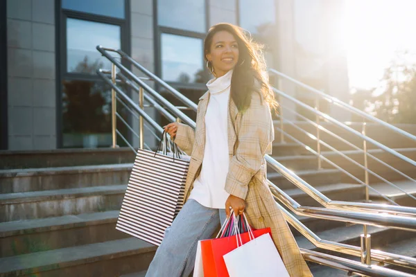 Stylish woman in a coat holds paper shopping bags, walks along the sunny streets of the city. Concept of shopping, shopping, lifestyle.