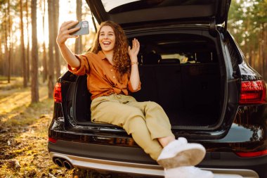 Genç bir kadın elinde telefonla arabanın bagajında gün batımına hayran. Kıvırcık saçlı bir turist cep telefonuyla fotoğraf çeker ve video aracılığıyla iletişim kurar. Seyahat, hafta sonu konsepti.