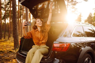Genç bir kadın elinde telefonla arabanın bagajında gün batımına hayran. Kıvırcık saçlı bir turist cep telefonuyla fotoğraf çeker ve video aracılığıyla iletişim kurar. Seyahat, hafta sonu konsepti.