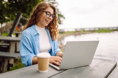 Parktaki bir masada oturan şık bir kadın gölün arka planında dizüstü bilgisayarlı ve sıcak bir içecekle oturuyor. Kahveli ve laptoplu kıvırcık kadın dışarıda güzel manzaralı çalışıyor..