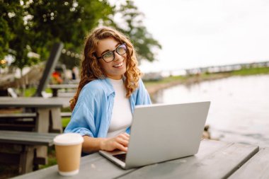 Parktaki bir masada oturan şık bir kadın gölün arka planında dizüstü bilgisayarlı ve sıcak bir içecekle oturuyor. Kahveli ve laptoplu kıvırcık kadın dışarıda güzel manzaralı çalışıyor..