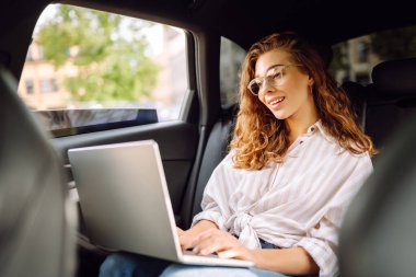 Dizüstü bilgisayarı olan şık bir kadın arabanın arka koltuğunda çalışıyor. İnternette çalışıyorum. İş seyahati. Teknoloji konsepti.