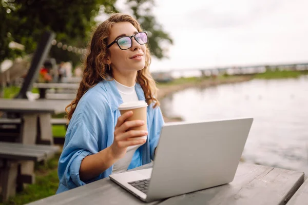 Stylish woman sitting at a table in a park against the backdrop of a lake with a laptop and a hot drink with her. Curly woman with coffee and laptop working outdoors with a beautiful view.