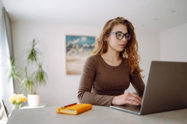 Günlük giysiler içindeki iş kadını dizüstü bilgisayar ve not defteriyle çalışıyor. Genç bir kadın müşterileriyle internet üzerinden görüşüyor, çalışıyor ve modern bir kuna üzerinde otururken ders çalışıyor. Eğitim kavramı, serbest çalışma.