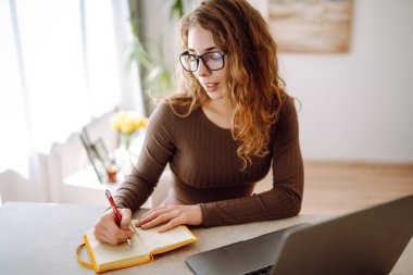 Günlük giysiler içindeki iş kadını dizüstü bilgisayar ve not defteriyle çalışıyor. Genç bir kadın müşterileriyle internet üzerinden görüşüyor, çalışıyor ve modern bir kuna üzerinde otururken ders çalışıyor. Eğitim kavramı, serbest çalışma.