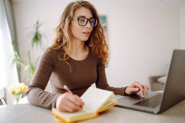 Günlük giysiler içindeki iş kadını dizüstü bilgisayar ve not defteriyle çalışıyor. Genç bir kadın müşterileriyle internet üzerinden görüşüyor, çalışıyor ve modern bir kuna üzerinde otururken ders çalışıyor. Eğitim kavramı, serbest çalışma.