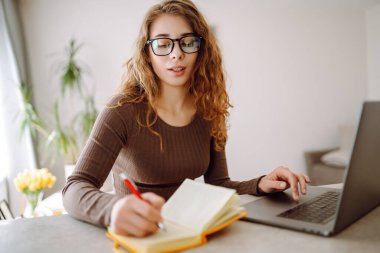 Günlük giysiler içindeki iş kadını dizüstü bilgisayar ve not defteriyle çalışıyor. Genç bir kadın müşterileriyle internet üzerinden görüşüyor, çalışıyor ve modern bir kuna üzerinde otururken ders çalışıyor. Eğitim kavramı, serbest çalışma.