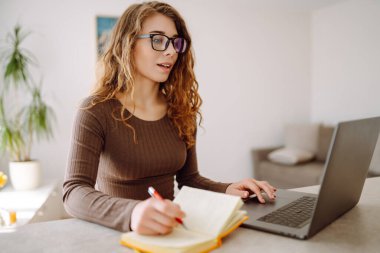 Günlük giysiler içindeki iş kadını dizüstü bilgisayar ve not defteriyle çalışıyor. Genç bir kadın müşterileriyle internet üzerinden görüşüyor, çalışıyor ve modern bir kuna üzerinde otururken ders çalışıyor. Eğitim kavramı, serbest çalışma.