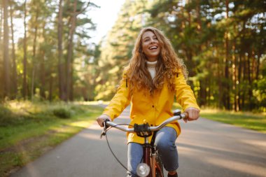 Sarı ceketli gülümseyen bir bayan turist bisiklet sürerken sonbahar parkında havanın tadını çıkarıyor. Sonbahar modası. Rahatlama kavramı, doğa..