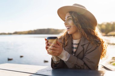Şapkalı neşeli bir kadın, gölün arka planında çay ya da kahve içer. Rahatlama, özgürlük kavramı..