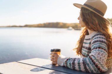 Şapkalı neşeli bir kadın, gölün arka planında çay ya da kahve içer. Rahatlama, özgürlük kavramı..