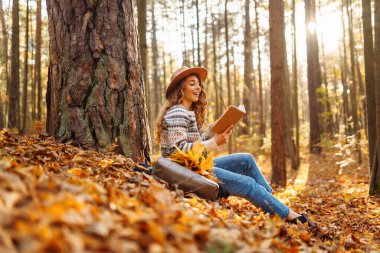 Şapkalı bir dişi turist, sonbahar ormanında sarı yaprakların üzerinde oturur ve kitap okur. Güzel kadın güneşli sonbahar havasının tadını çıkarıyor..