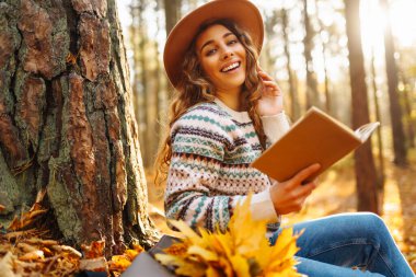 Şapkalı bir dişi turist, sonbahar ormanında sarı yaprakların üzerinde oturur ve kitap okur. Güzel kadın güneşli sonbahar havasının tadını çıkarıyor..