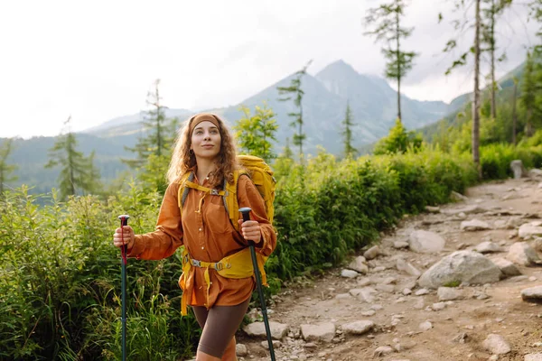 Young woman traveler with a yellow hiking backpack and hiking poles on trail among the mountains. Adventure concept, hiking. Active lifestyle.