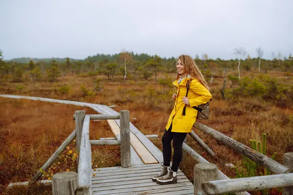 stock image A hiker female wearing a yellow coat and backpack walks along a scenic nature trail with a wooden boardwalk overlooking wetlands. Travel and exploration