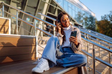 Kulaklık takan ve elinde telefon olan şık bir kadın açık havada şehir atmosferinin tadını çıkarıyor, müzik dinliyor, video dersleri alıyor. Sosyal ağlarda iletişim.