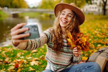 Şapkalı neşeli bir kadın elinde telefonla sonbahar parkında bir battaniyeye oturur, selfie çeker, video görüşmesiyle konuşur, bloglar yazar. Doğadan zevk alma kavramı.