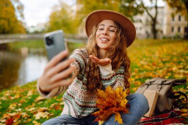 Şapkalı neşeli bir kadın elinde telefonla sonbahar parkında bir battaniyeye oturur, selfie çeker, video görüşmesiyle konuşur, bloglar yazar. Doğadan zevk alma kavramı.