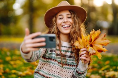 Şapkalı neşeli bir kadın elinde telefonla sonbahar parkında bir battaniyeye oturur, selfie çeker, video görüşmesiyle konuşur, bloglar yazar. Doğadan zevk alma kavramı.