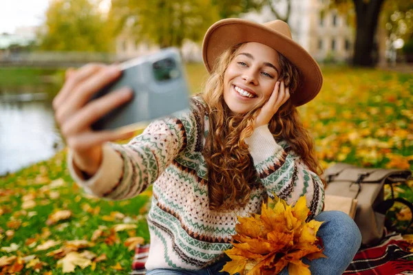 Şapkalı neşeli bir kadın elinde telefonla sonbahar parkında bir battaniyeye oturur, selfie çeker, video görüşmesiyle konuşur, bloglar yazar. Doğadan zevk alma kavramı.
