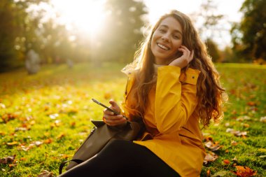 Gülümseyen bir kadın elinde telefonuyla bahçedeki sarı sonbahar yapraklarının arasında oturuyor. Bir dişi sonbahar parkında akıllı telefon kullanıyor, doğanın tadını çıkarıyor..