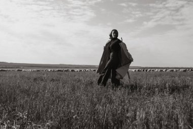 Tarlada, kuru otların arasında paltolu şık bir kadın, doğanın tadını çıkarıyor. Siyah beyaz fotoğraf. Doğa, moda, özgürlük kavramı. Yaşam biçimi.