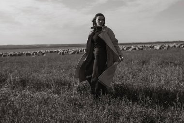Tarlada, kuru otların arasında paltolu şık bir kadın, doğanın tadını çıkarıyor. Siyah beyaz fotoğraf. Doğa, moda, özgürlük kavramı. Yaşam biçimi.
