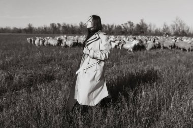Tarlada, kuru otların arasında paltolu şık bir kadın, doğanın tadını çıkarıyor. Siyah beyaz fotoğraf. Doğa, moda, özgürlük kavramı. Yaşam biçimi.