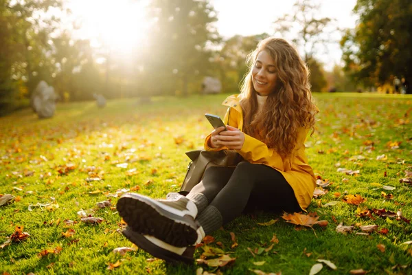 stock image A smiling woman sits outdoors on the lawn among yellow autumn leaves with a phone in her hands. A female uses a smartphone in an autumn park, enjoying nature.