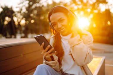 Şık günlük kıyafetler ve kablosuz kulaklıklar takan esmer kadın gün batımında şehirde otururken elinde bir telefon tutuyor. Güzel bir kadın havanın tadını çıkarıyor ve dışarıda müzik dinliyor..