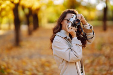 Şık giyinmiş mutlu genç bir kadın eski moda bir kamerayla fotoğraf çekiyor, sonbahar güneşli parkında eğleniyor ve dinleniyor. Rahatlama konsepti, boş zaman aktiviteleri.