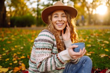 Şapkalı rahat bir kadın, parktaki güneşli havanın tadını çıkarıyor, kırmızı bir battaniyenin üzerinde oturuyor, termostan sıcak bir içecek içiyor. Dinlenme, rahatlama kavramı. Yaşam biçimi.
