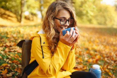 Sarı yağmurluklu güzel bir turist sarı yaprakların arasında bir açıklıkta oturur, termostan çay içer, sonbahar parkında güneşli havanın tadını çıkarır.. 