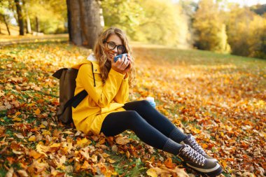 Sarı yağmurluklu güzel bir turist sarı yaprakların arasında bir açıklıkta oturur, termostan çay içer, sonbahar parkında güneşli havanın tadını çıkarır.. 