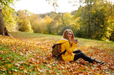 Sarı yağmurluklu güzel bir turist sarı yaprakların arasında bir açıklıkta oturur, termostan çay içer, sonbahar parkında güneşli havanın tadını çıkarır.. 