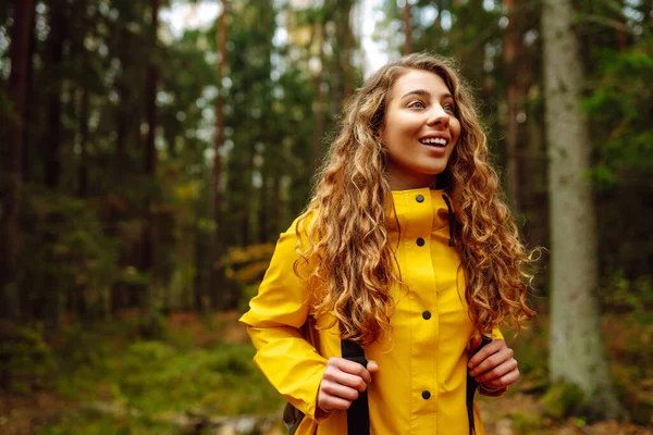 黄色いコートの幸せな女性は 彼女の周りの緑の美しい自然の森を楽しんでいます バックパック付きの観光客は 森のチケットを歩いて新しい場所を探索しています — ストック写真