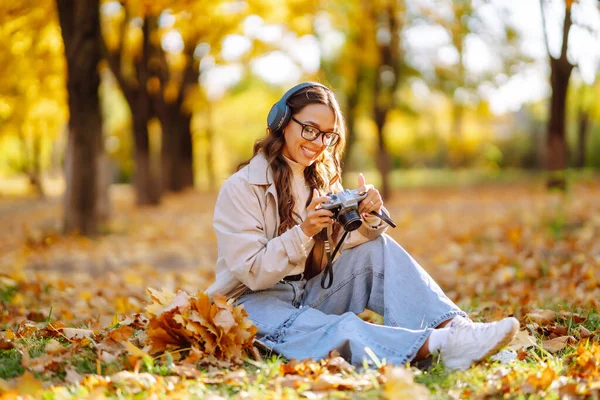 stock image Happy young woman in stylish clothes takes pictures with a retro camera, having fun and relaxing in the autumn sunny park. The concept of relaxation, leisure activities.