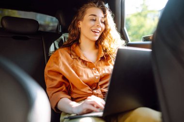 Dizüstü bilgisayarı olan genç, kıvırcık bir kadın arabanın arka koltuğunda oturuyor. Güzel bir kadın arabada dizüstü bilgisayar kullanır. Uzak çalışma, serbest çalışma.