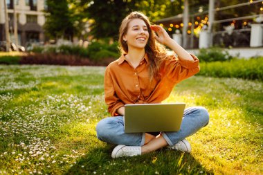 Dışarıda dizüstü bilgisayarı olan neşeli bir kadın. Genç bir kadın yeşil çimlerde oturuyor ve dizüstü bilgisayarla çalışıyor. Blog yazma kavramı, serbest çalışma, rahatla.
