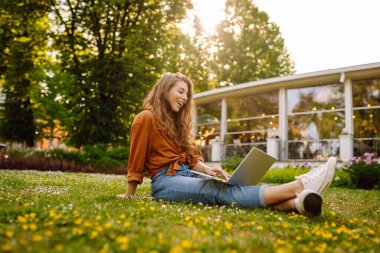 Dışarıda dizüstü bilgisayarı olan neşeli bir kadın. Genç bir kadın yeşil çimlerde oturuyor ve dizüstü bilgisayarla çalışıyor. Blog yazma kavramı, serbest çalışma, rahatla.