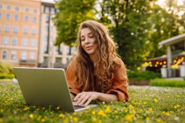 Dışarıda dizüstü bilgisayarı olan neşeli bir kadın. Genç bir kadın yeşil çimlerde oturuyor ve dizüstü bilgisayarla çalışıyor. Blog yazma kavramı, serbest çalışma, rahatla.