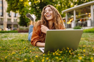 Dışarıda dizüstü bilgisayarı olan neşeli bir kadın. Genç bir kadın yeşil çimlerde oturuyor ve dizüstü bilgisayarla çalışıyor. Blog yazma kavramı, serbest çalışma, rahatla.