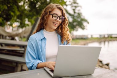 Göl kenarındaki bir masada dizüstü bilgisayarla oturan günlük kıyafetli neşeli bir kadın. Dışarıda dizüstü bilgisayarla çalışan hoş bir kadın. Serbest meslek, teknoloji konsepti.