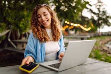 Göl kenarındaki bir masada dizüstü bilgisayarla oturan günlük kıyafetli neşeli bir kadın. Dışarıda dizüstü bilgisayarla çalışan hoş bir kadın. Serbest meslek, teknoloji konsepti.