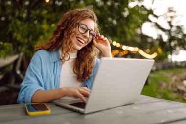 Göl kenarındaki bir masada dizüstü bilgisayarla oturan günlük kıyafetli neşeli bir kadın. Dışarıda dizüstü bilgisayarla çalışan hoş bir kadın. Serbest meslek, teknoloji konsepti.