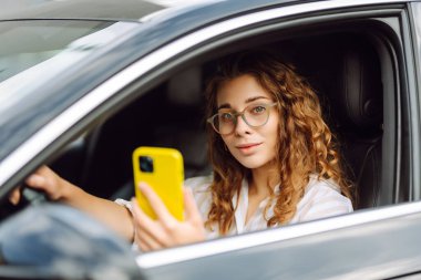 Direksiyonun başında oturan genç ve şık bir kadın akıllı telefon kullanıyor. Bir kadın sürücü harita çıkarmak ya da park ücretini ödemek için bir cep telefonu kullanır. Teknoloji konsepti.
