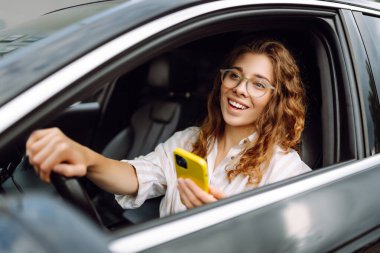 Direksiyonun başında oturan genç ve şık bir kadın akıllı telefon kullanıyor. Bir kadın sürücü harita çıkarmak ya da park ücretini ödemek için bir cep telefonu kullanır. Teknoloji konsepti.