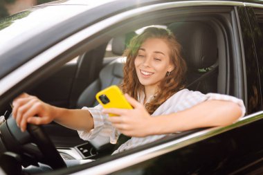 Direksiyonun başında oturan genç ve şık bir kadın akıllı telefon kullanıyor. Bir kadın sürücü harita çıkarmak ya da park ücretini ödemek için bir cep telefonu kullanır. Teknoloji konsepti.