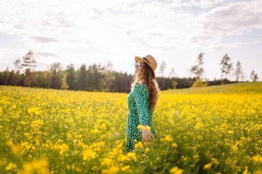 Mutlu kadın eğleniyor ve kolza tohumu tarlasında yürüyor. Parlak elbiseli ve şapkalı güzel bir kadın güneşli havanın tadını çıkarıyor. Doğa kavramı, rahatlama..