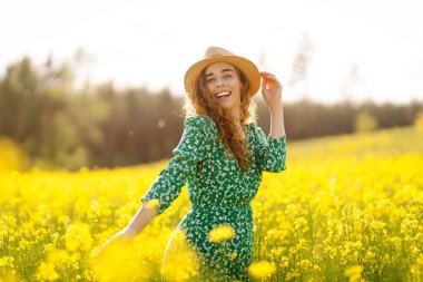 Mutlu kadın eğleniyor ve kolza tohumu tarlasında yürüyor. Parlak elbiseli ve şapkalı güzel bir kadın güneşli havanın tadını çıkarıyor. Doğa kavramı, rahatlama..